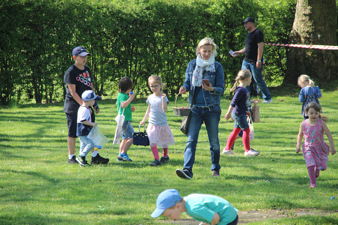 Familie und Kinder spielen im Freien auf einer Wiese, umgeben von B&auml;umen und farbenfrohen Kleidungen.