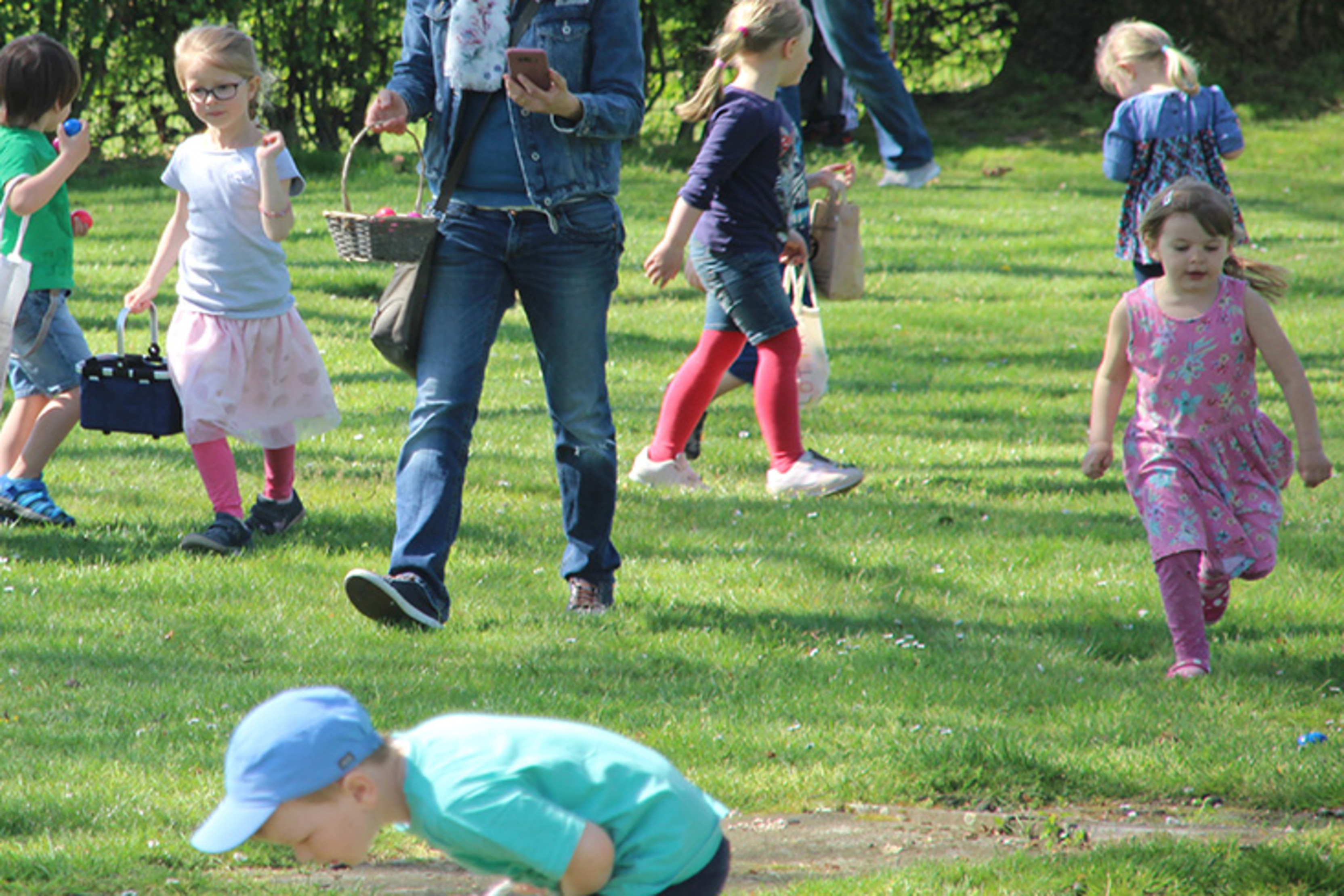 Familie und Kinder spielen im Freien auf einer Wiese, umgeben von B&auml;umen und farbenfrohen Kleidungen.