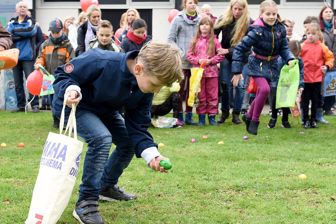Kind hebt ein buntes Plastik-Ei auf, w&auml;hrend andere Kinder und Erwachsene im Hintergrund auf einer Wiese stehen.
