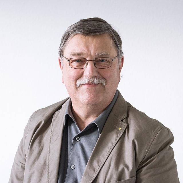 Porträt eines älteren Mannes mit Brille, lächelnd, in einem hellen Anzug und Hemd, vor einem neutralen Hintergrund.