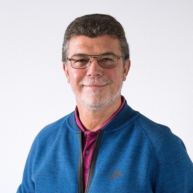 Mann mit Brille und grauem Haar l&auml;chelt in blauer Jacke und rotem Polo-Shirt vor neutralem Hintergrund.