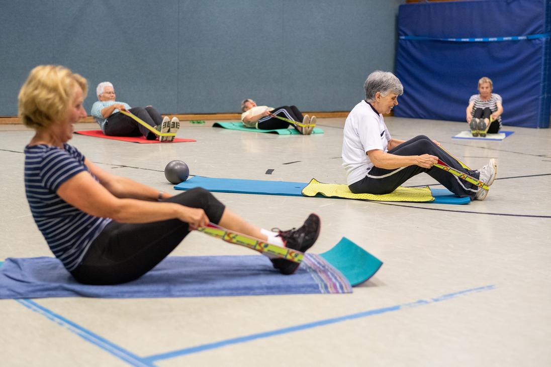 Senioren führen Rückenübungen mit Widerstandsbändern auf Matten in einer Sporthalle durch. Diese sind in verschiedenen Farben.