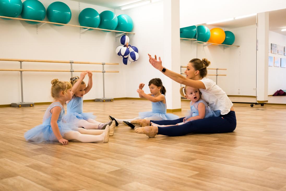 Tanzlehrerin sitzt auf dem Boden mit vier Vorschulmädchen in blauen Tutus in einem modernen Tanzraum.