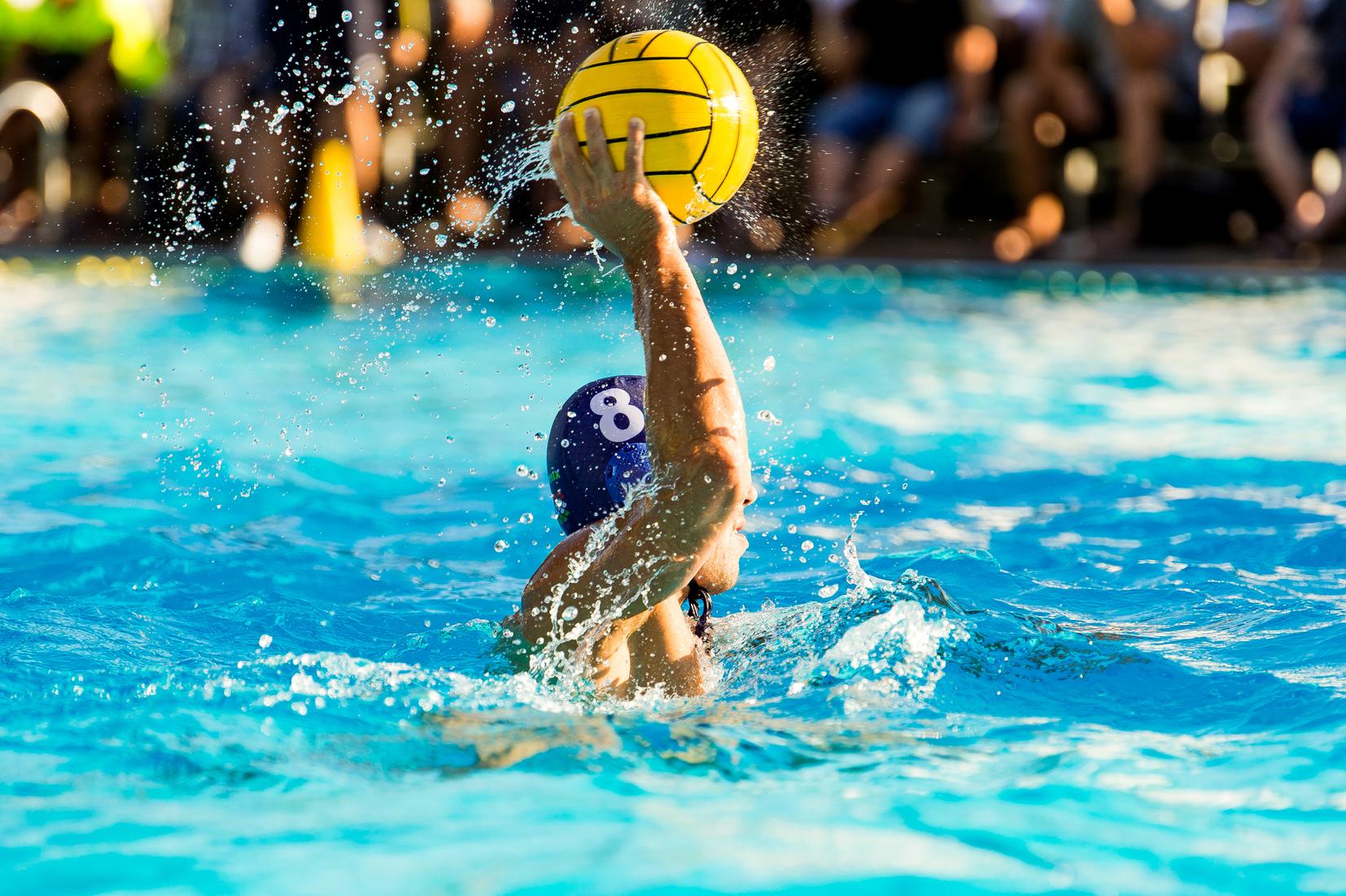 Wasserspieler im blauer Badekappe wirft einen gelben Wasserball aus dem Wasser, spritzendes Wasser um ihn herum.