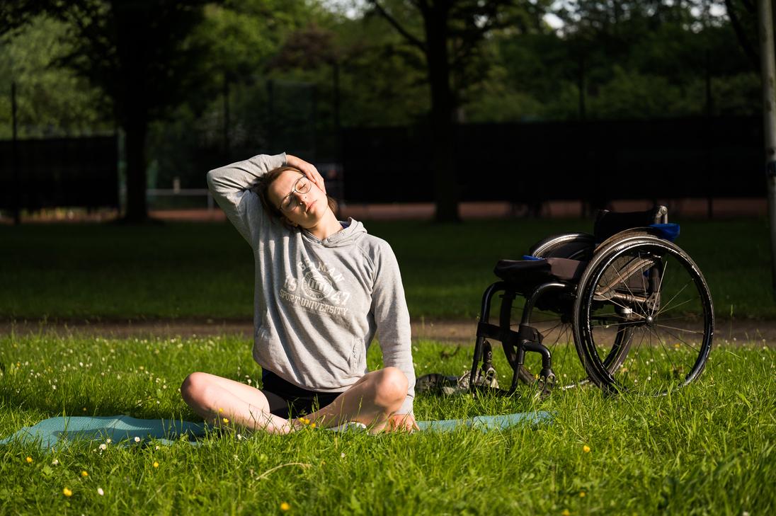 Junge Frau macht Nackenübung auf einer Yogamatte im Freien, neben ihr steht ein Rollstuhl.